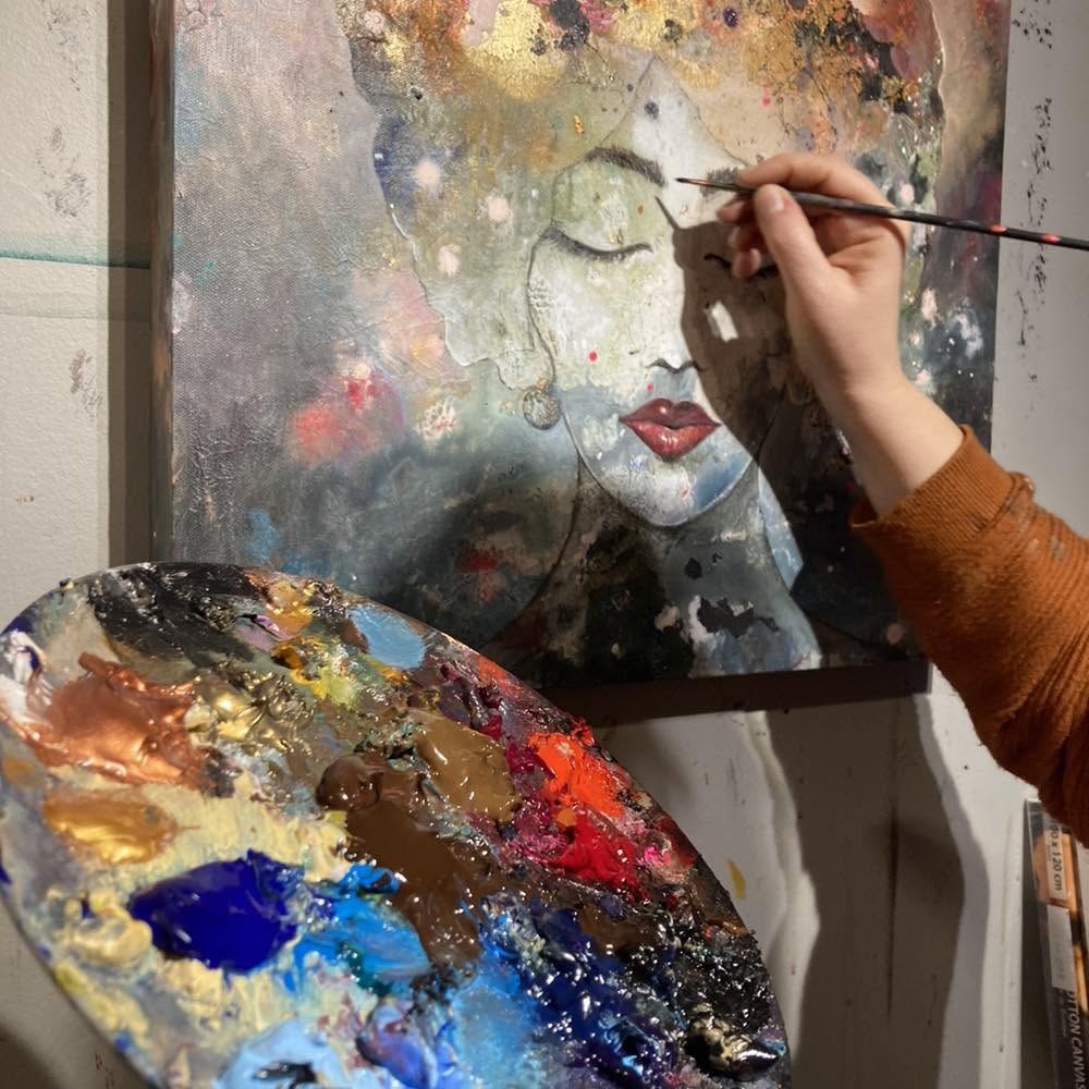 Carmen's hand holding a fine brush over a portrait in progress, a paint-covered palette in the foreground
