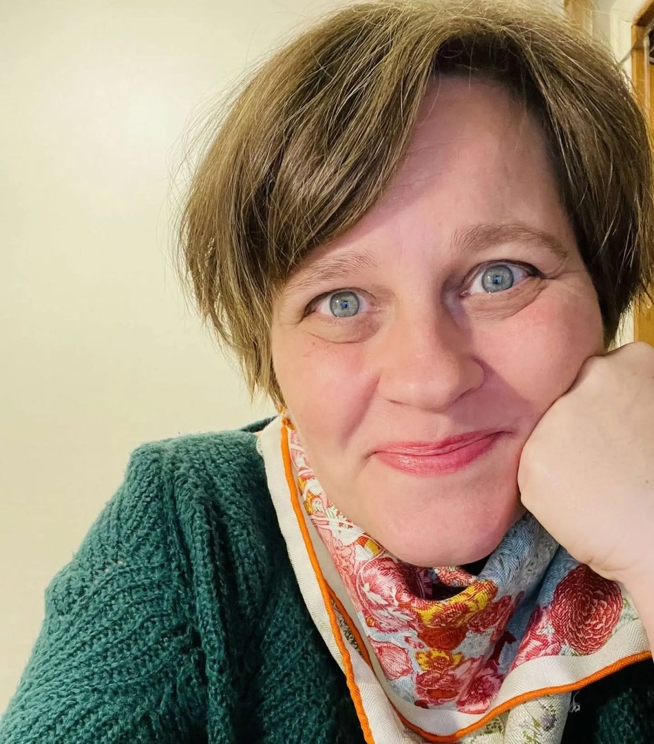 Carmen Faunbäck in a green sweater and a patterned silk scarf, resting her chin on her hand and smiling warmly at the camera